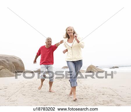 Stock Image - Senior Couple On Holiday Running Along Winter Beach. Fotosearch