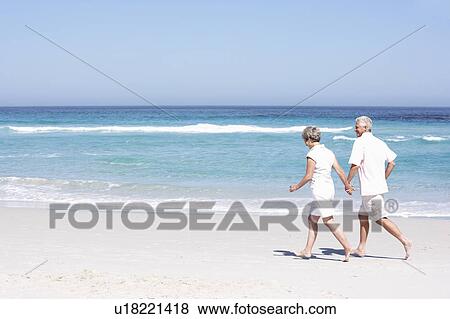 Stock Photo - Senior Couple On Holiday Running Along Sandy Beach. Fotosearch