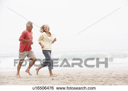 Stock Photograph - Senior Couple On Holiday Running Along Winter Beach. Fotosearch