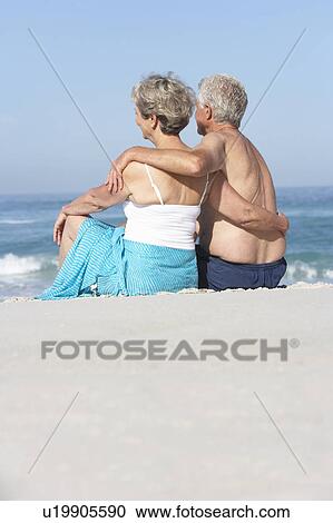 Stock Image - Senior Couple On Holiday Sitting On Sandy Beach. Fotosearch