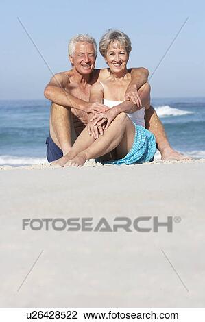 Stock Image - Senior Couple On Holiday Sitting On Sandy Beach. Fotosearch