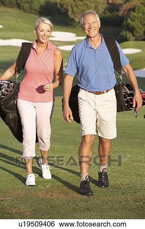 Stock Photograph - Senior Couple Walking Along Golf Course Carrying Bags. Fotosearch
