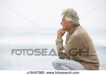 Stock Image - Senior Man On Holiday Kneeling On Winter Beach. Fotosearch