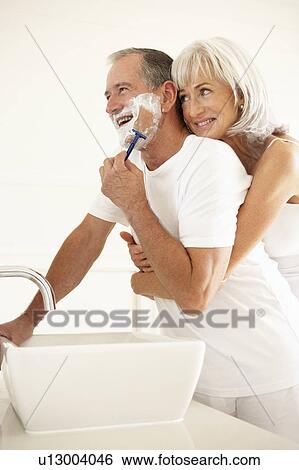 Stock Photograph - Senior Man Shaving In Bathroom Mirror With Wife Watching. Fotosearch