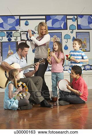 Stock Image - Teachers Playing Guitar With Pupils Having Music Lesson In Classroom. Fotosearch