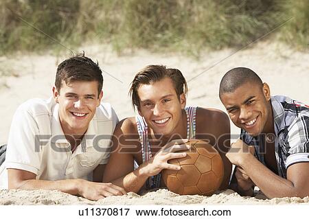 Stock Photo - Three Young Men Relaxing On Beach With Football Together. Fotosearch