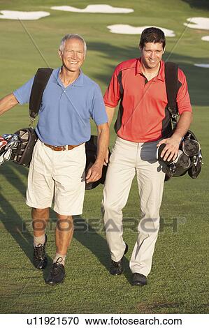 Stock Image - Two Men Walking Along Golf Course Carrying Bags. Fotosearch