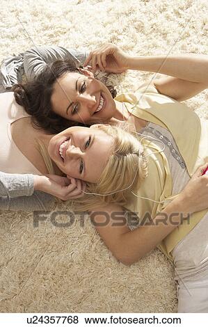 Stock Photo - Two Women Listening To MP3 Player On Headphones Together Relaxing Laying On Rug At Home . Fotosearch