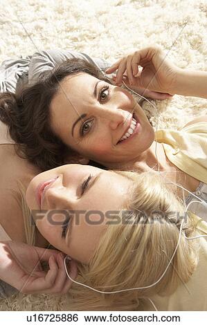 Stock Photograph - Two Women Listening To MP3 Player On Headphones Together Relaxing Laying On Rug At Home . Fotosearch
