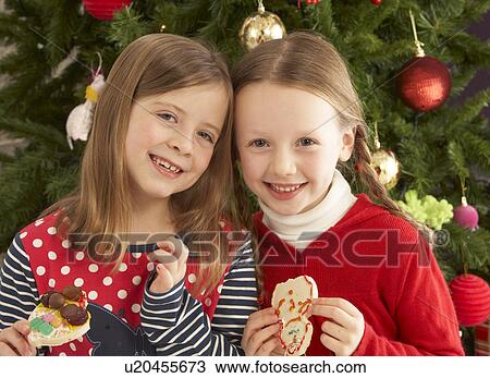 Stock Image - Two Young Girls Eating Cookies In Front Of Christmas Tree. Fotosearch