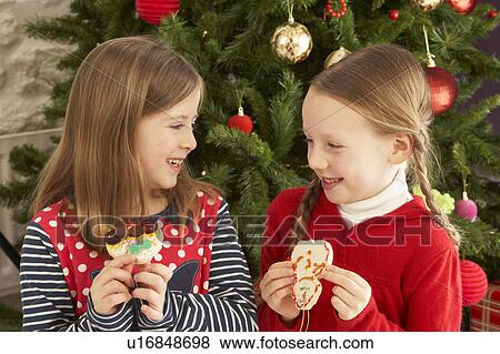 Stock Photo - Two Young Girls Eating Cookies In Front Of Christmas Tree. Fotosearch