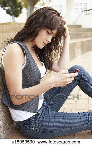 Stock Image - Unhappy Female Teenage Student Sitting Outside On College Steps Using Mobile Phone. Fotosearch