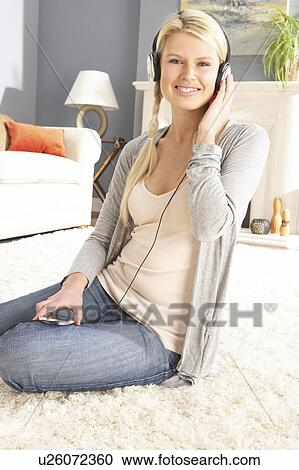 Stock Image - Woman Listening To MP3 Player On Headphones Relaxing Sitting On Rug At Home . Fotosearch