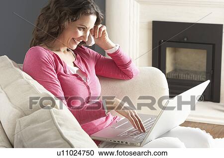 Stock Image - Woman Using Laptop Relaxing Sitting On Sofa At Home Talking On Phone. Fotosearch