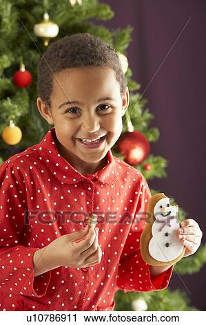 Stock Image - Young Boy Eating Cookie In Front Of Christmas Tree. Fotosearch