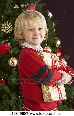 Stock Image - Young Boy Holding Gift In Front Of Christmas Tree. Fotosearch