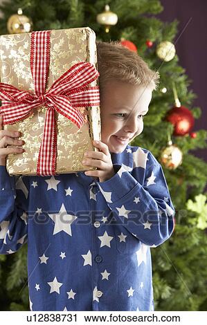 Stock Image - Young Boy Holding Wrapped Present In Front Of Christmas Tree. Fotosearch