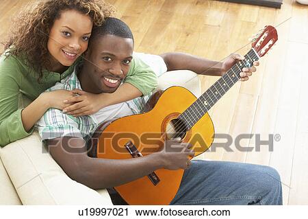 Stock Image - Young Couple Relaxing Sitting On Sofa Playing Acoustic Guitar. Fotosearch