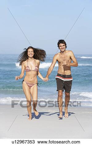 Stock Image - Young Couple Running On Beach Wearing Swimwear. Fotosearch