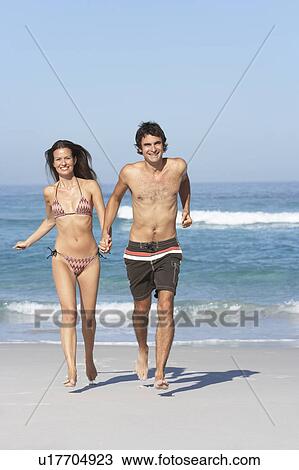 Stock Image - Young Couple Running On Beach Wearing Swimwear. Fotosearch