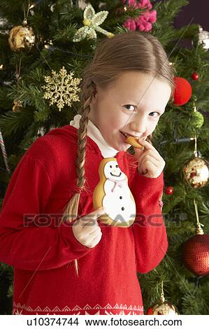 Picture - Young Girl Eating Cookie In Front Of Christmas Tree. Fotosearch