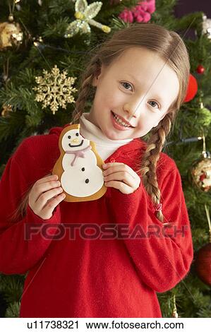 Stock Image - Young Girl Eating Cookie In Front Of Christmas Tree. Fotosearch