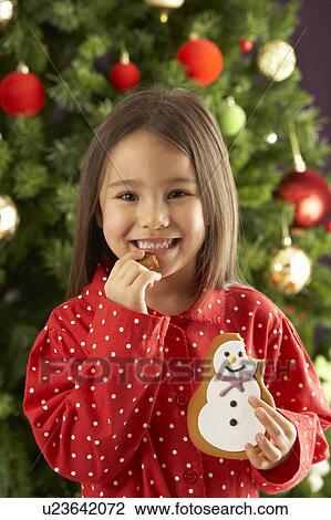 Stock Image - Young Girl Eating Cookie In Front Of Christmas Tree. Fotosearch