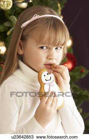 Stock Image - Young Girl Eating Cookie In Front Of Christmas Tree. Fotosearch