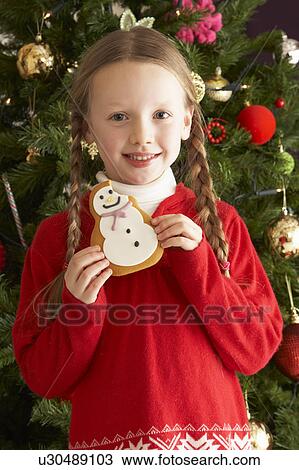 Stock Image - Young Girl Eating Cookie In Front Of Christmas Tree. Fotosearch