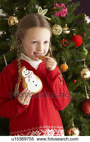 Stock Photography - Young Girl Eating Cookie In Front Of Christmas Tree. Fotosearch