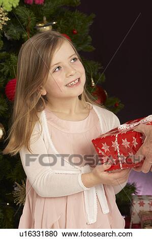 Stock Image - Young Girl Holding Christmas Present In Front Of Christmas Tree. Fotosearch