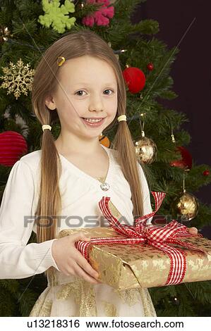 Stock Photograph - Young Girl Holding Christmas Present In Front Of Christmas Tree. Fotosearch