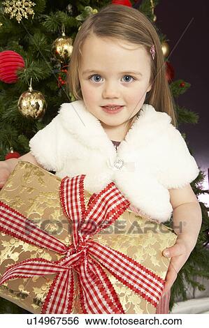 Stock Photograph - Young Girl Holding Gift In Front Of Christmas Tree. Fotosearch