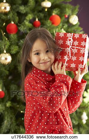 Stock Photograph - Young Girl Holding Wrapped Present In Front Of Christmas Tree. Fotosearch