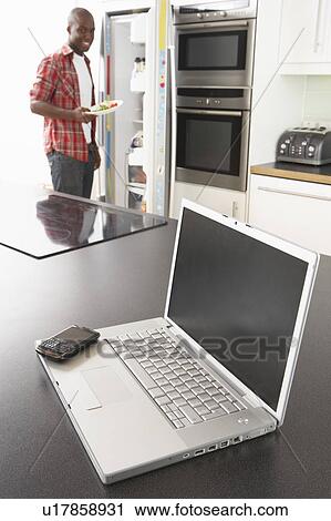 Stock Image - Young Man Fixing Snack In Kitchen With Laptop In Modern Kitchen . Fotosearch