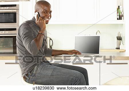 Stock Image - Young Man Relaxing Sitting In Kitchen Talking On Phone. Fotosearch