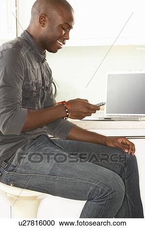 Stock Image - Young Man Relaxing Sitting In Kitchen Talking On Phone. Fotosearch