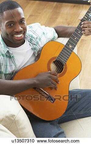 Stock Image - Young Man Relaxing Sitting On Sofa Playing Acoustic Guitar. Fotosearch
