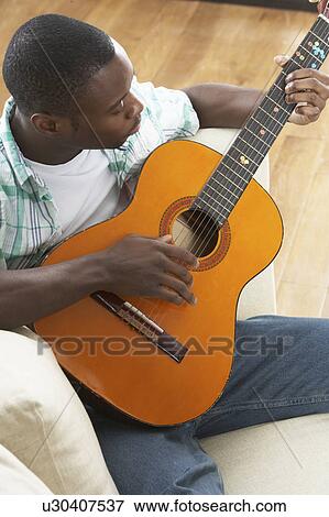 Stock Photo - Young Man Relaxing Sitting On Sofa Playing Acoustic Guitar. Fotosearch