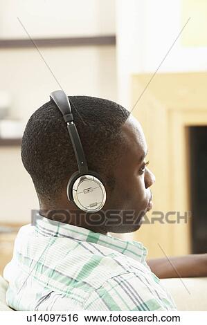 Stock Photograph - Young Man Relaxing Sitting On Sofa Listening to Music At Home On Headphones. Fotosearch