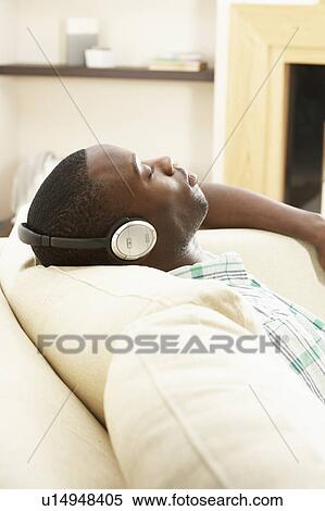 Stock Photography - Young Man Relaxing Sitting On Sofa Listening to Music At Home On Headphones. Fotosearch