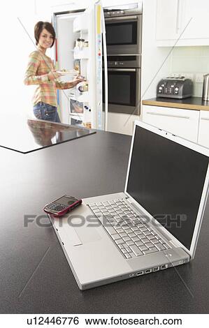 Stock Photograph - Young Woman Fixing Snack In Kitchen With Laptop In Modern Kitchen . Fotosearch