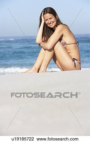 Stock Image - Young Woman Sitting On Sandy Beach On Holiday Wearing Bikini. Fotosearch