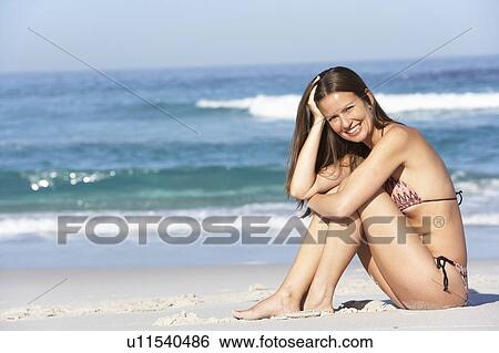 Stock Photograph - Young Woman Sitting On Sandy Beach On Holiday Wearing Bikini. Fotosearch