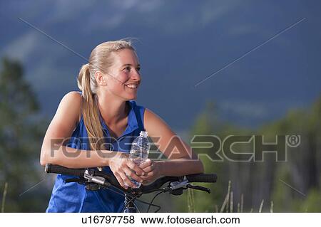 Canada, British Columbia, Fernie, Mid adult woman leaning against bike handlebar and drinking water View Large Photo Image Stock Photo - Canada, British Columbia, Fernie, Mid adult woman leaning against bike handlebar and drinking water. Fotosearch