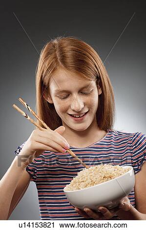 Stock Image - Portrait of redhead girl (10-11) holding chopsticks and bowl of rice, studio shot. Fotosearch