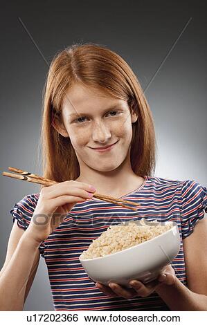 Stock Photograph - Portrait of redhead girl (10-11) holding chopsticks and bowl of rice, studio shot. Fotosearch