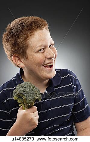 Stock Image - Portrait of redhead teenage boy (14-15) holding broccoli and laughing, studio shot. Fotosearch