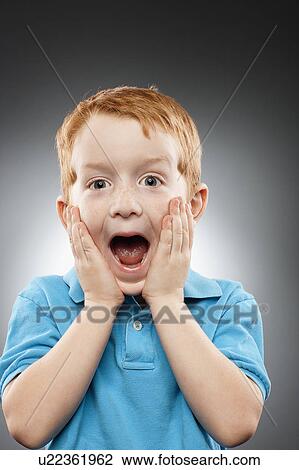 Stock Image - Portrait of smiling redhead boy (4-5) wearing blue polo shirt and looking surprised, studio shot. Fotosearch