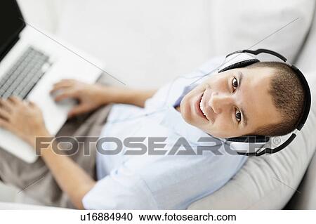 Stock Image - South Africa, Young man wearing headphones sitting on sofa and using laptop, high angle view. Fotosearch